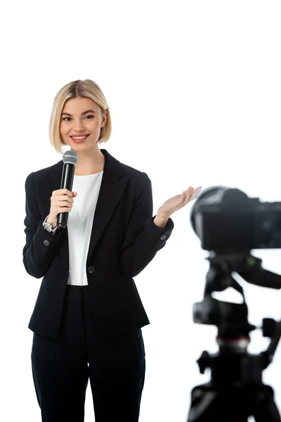 Lächelnder blonder Kommentator mit Mikrofon, das mit der Hand in der Nähe der Digitalkamera auf unscharfem, auf weiß isoliertem Vordergrund zeigt — Stockfoto