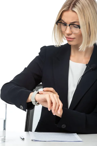 Frowning anchorwoman looking at wristwatch while sitting at workplace isolated on white — Stock Photo