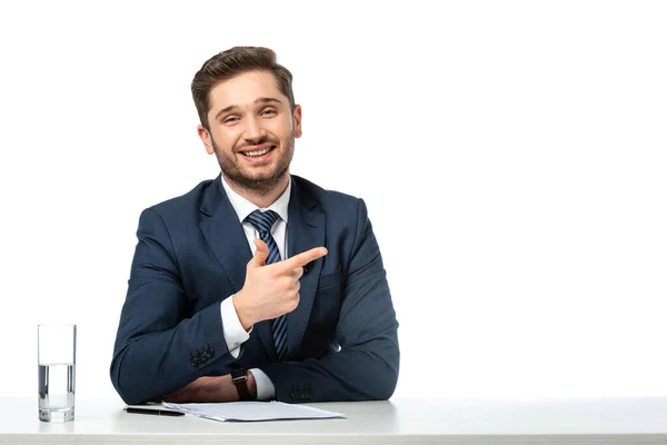 Feliz presentador señalando a un lado con el dedo cerca de los papeles y el vaso de agua aislado en blanco - foto de stock