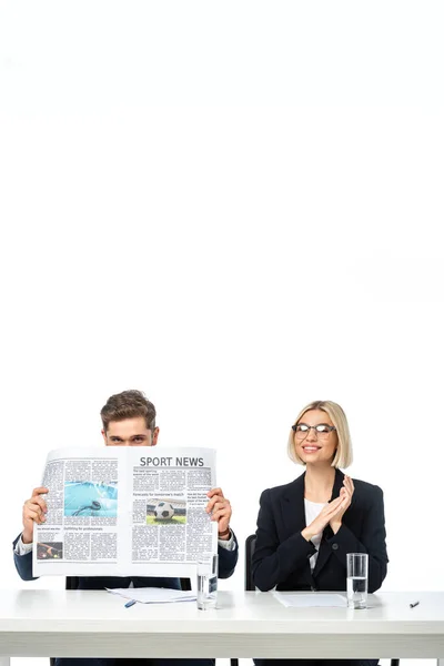 Alegre periodista rubia sonriendo cerca de colega oscureciendo la cara con periódico aislado en blanco - foto de stock