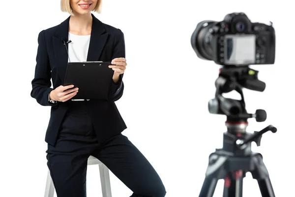 Vista recortada de presentadora sonriente con portapapeles sentado cerca de la cámara digital en primer plano borroso aislado en blanco - foto de stock