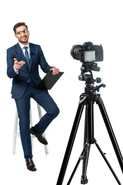 Fröhlicher Nachrichtenanker, der mit der Hand zeigt, während er auf einem hohen Hocker neben der Digitalkamera auf weißem, verschwommenem Vordergrund sitzt — Stockfoto