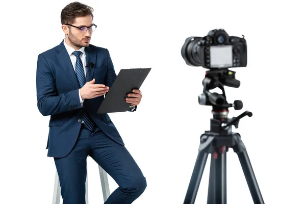 Nachrichtenanker, der auf Klemmbrett zeigt, während er auf einem hohen Hocker in der Nähe der Digitalkamera auf unscharfem Vordergrund sitzt, isoliert auf Weiß — Stockfoto