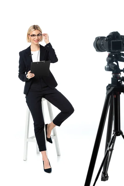 Lächelnder Nachrichtensprecher, der Brillen berührt, während er auf einem hohen Hocker in der Nähe der Digitalkamera auf weiß sitzt — Stockfoto