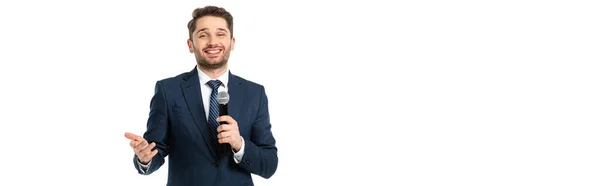 Cheerful journalist with microphone pointing with hand while smiling at camera isolated on white, banner — Stock Photo