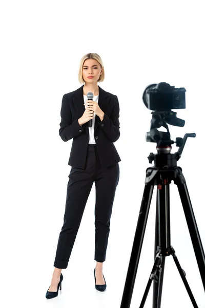 Full length view of blonde news anchor with microphone near digital camera on white, blurred foreground — Stock Photo
