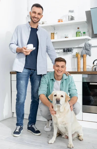Щасливий чоловік тримає чашку і стоїть біля чоловіка і собаки на кухні — стокове фото