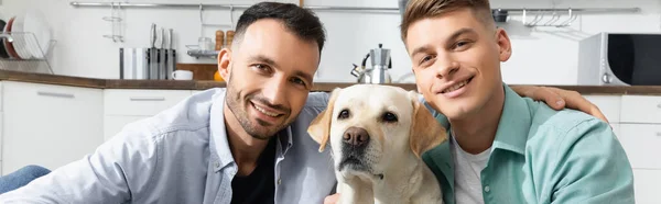 Веселі гомосексуальні чоловіки посміхаються з собакою вдома, банер — стокове фото