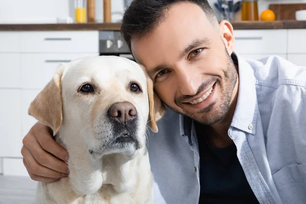 Hombre barbudo alegre sonriendo cerca de labrador en casa - foto de stock