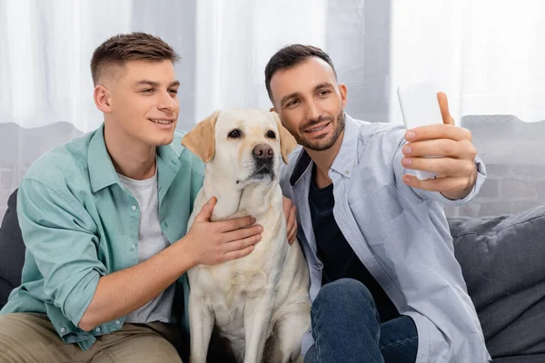 Alegre mismo sexo pareja sonriendo y tomando selfie con labrador - foto de stock