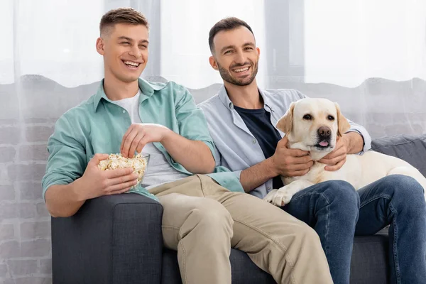 Cheerful same sex couple smiling and watching movie with popcorn and labrador — Stock Photo