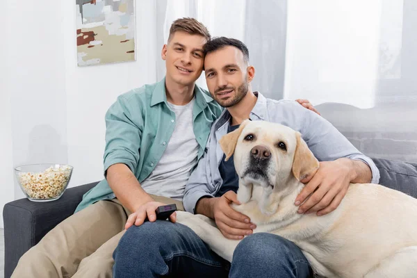 Alegre pareja del mismo sexo sonriendo cerca de bowl con palomitas de maíz y labrador — Stock Photo