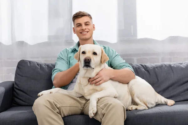 Alegre homem sentado no sofá e abraçando labrador — Fotografia de Stock