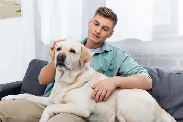 Homem satisfeito sentado no sofá e abraçando labrador — Fotografia de Stock