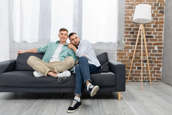 Fröhliches gleichgeschlechtliches Paar schaut weg, während es zu Hause auf der Couch sitzt — Stockfoto
