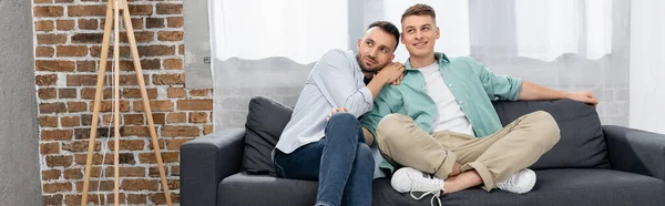 Fröhliches gleichgeschlechtliches Paar, das wegschaut, während es zu Hause auf der Couch sitzt, Banner — Stockfoto