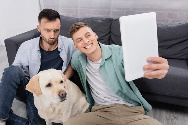 Happy same sex couple taking selfie on digital tablet near labrador in living room — Stock Photo