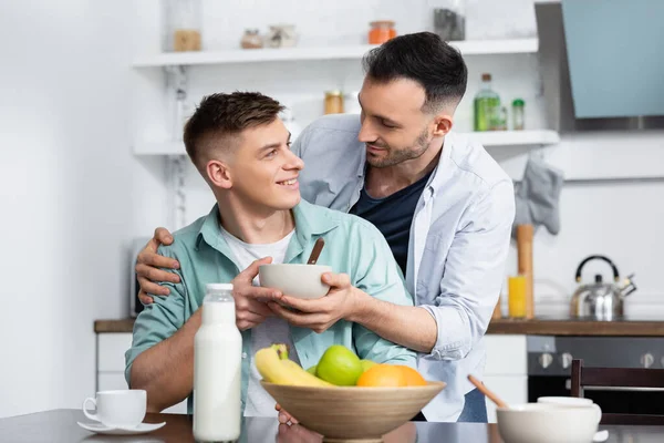 Hombre homosexual sosteniendo tazón cerca de marido y frutas en la mesa - foto de stock