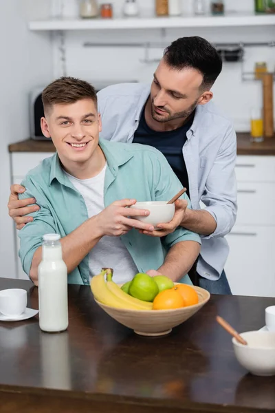 Гомосексуальный мужчина держит чашу рядом счастливый муж и фрукты на столе — стоковое фото