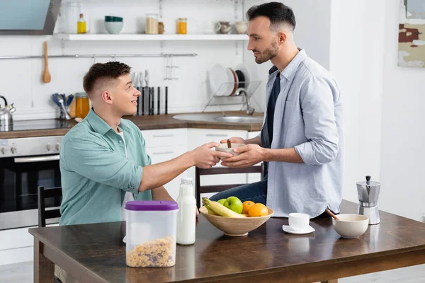 Homosexual man giving bowl to happy husband near fruits on table — Stock Photo