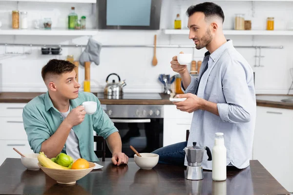 Happy same sex couple looking at each other while holding cups in kitchen — Stock Photo