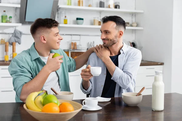 Hombre homosexual feliz sosteniendo la taza y mirando al marido en la cocina - foto de stock