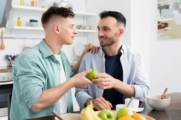 Heureux homosexuel homme donnant pomme à mari dans la cuisine — Photo de stock