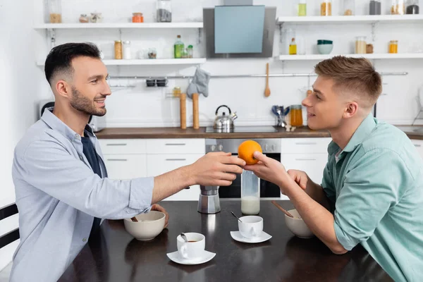 Side view of happy homosexual man giving orange to husband during breakfast — Stock Photo