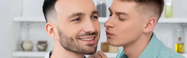 Feliz hombre haciendo pucheros labios mientras besa mejilla de sonriente marido, bandera - foto de stock