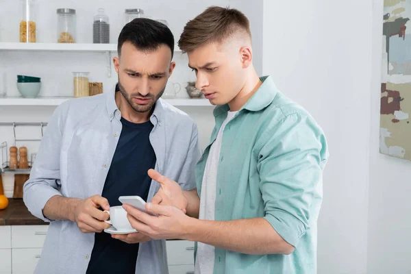 Homosexuel homme pointant avec la main à smartphone près de mari avec tasse de café — Photo de stock