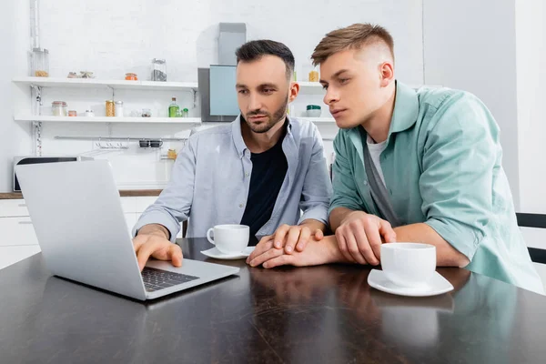 Гомосексуальная пара смотрит на ноутбук возле чашек на столе — стоковое фото