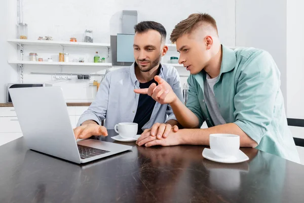 Uomo omosessuale che punta con il dito e guardando il computer portatile vicino al marito e tazze sul tavolo — Foto stock