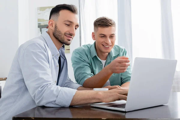 Homosexual man pointing with finger and looking at laptop near happy husband — Stock Photo