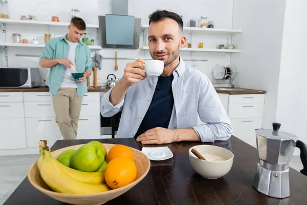 Feliz homosexual hombre beber café cerca marido en borrosa fondo - foto de stock