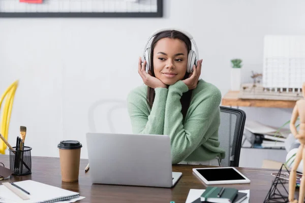 Heureux designer d'intérieur afro-américain dans les écouteurs assis sur le lieu de travail, flou au premier plan — Photo de stock