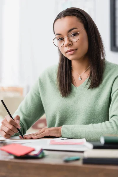 Designer d'intérieur afro-américain en lunettes regardant la caméra tout en tenant un crayon sur le lieu de travail, premier plan flou — Photo de stock