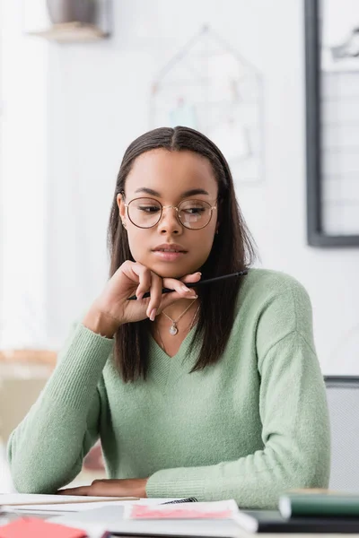 Designer d'intérieur afro-américain rêveur dans des lunettes tenant stylo tout en travaillant à la maison — Photo de stock