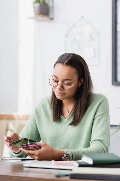 Sorridente interior designer africano americano che tiene campioni di materiale sul posto di lavoro — Foto stock