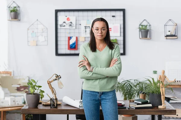 Selbstbewusster afrikanisch-amerikanischer Architekt steht mit verschränkten Armen in der Nähe seines Arbeitsplatzes und blickt in die Kamera — Stockfoto