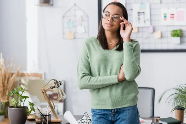 Junger afrikanisch-amerikanischer Architekt justiert Brille, während er in der Nähe von Schreibtisch wegschaut — Stockfoto