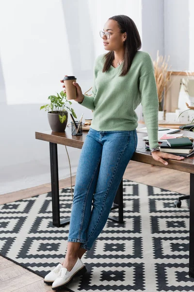 Junge afrikanisch-amerikanische Innenarchitektin schaut weg, während sie mit Kaffee in der Nähe des Schreibtisches steht — Stockfoto