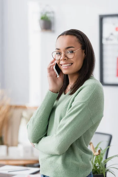 Sonriente diseñador de interiores afroamericano mirando a la cámara mientras habla por teléfono móvil - foto de stock