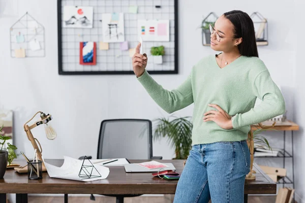 Joven diseñador de interiores afroamericano tomando selfie con la mano en la cadera en el estudio en casa - foto de stock