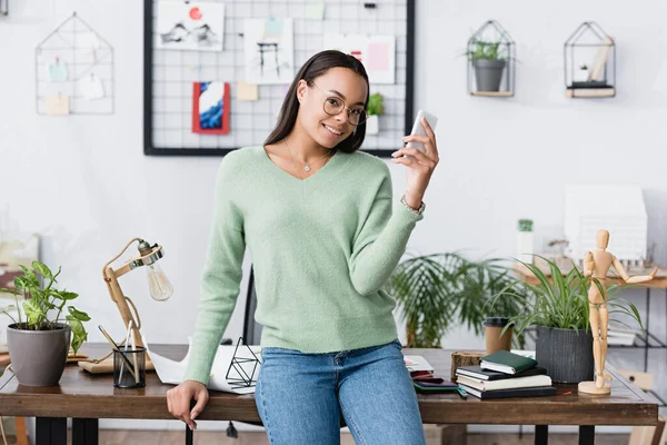 Heureux architecte afro-américain souriant à la caméra tandis que la messagerie sur smartphone — Photo de stock