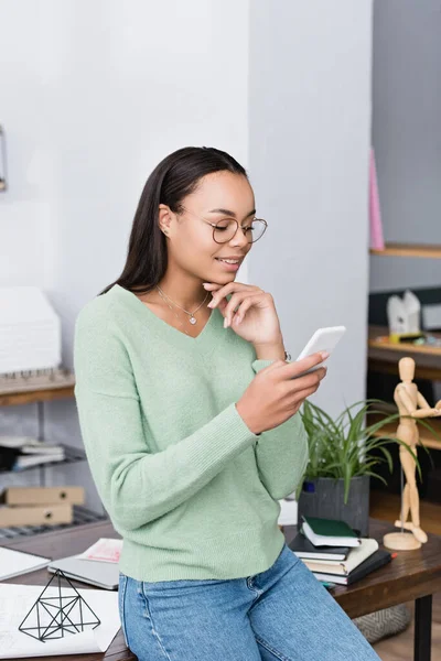 Sonriente afroamericano arquitecto mensajería en el teléfono inteligente cerca del lugar de trabajo en casa - foto de stock