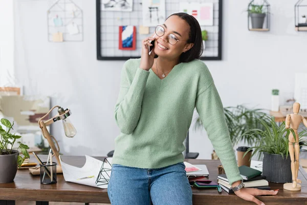 Cheerful african american interior designer talking on smartphone near desk at home — Stock Photo