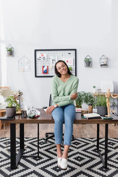 Smiling african american interior designer sitting on desk at home studio and looking at camera — Stock Photo