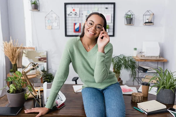 Happy african american architect adjusting eyeglasses while sitting on desk and smiling at camera — Stock Photo