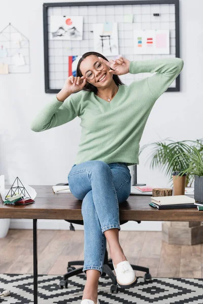 Alegre arquitecto afroamericano tocando gafas mientras está sentado en el escritorio en casa - foto de stock