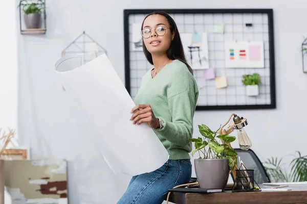 Young african american interior designer looking away while holding project near desk at home — Stock Photo